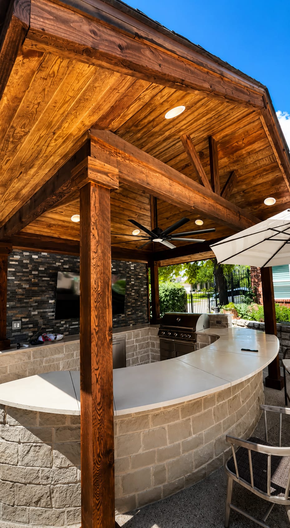 Curved stone outdoor kitchen bar under a stained cedar ceiling with recessed lights and a ceiling fan